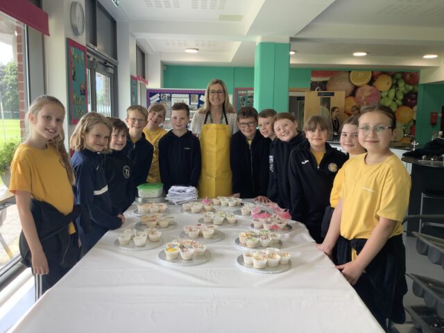 Pupils from a visiting school smiling with a Trent College teacher around a table of cakes they have made.